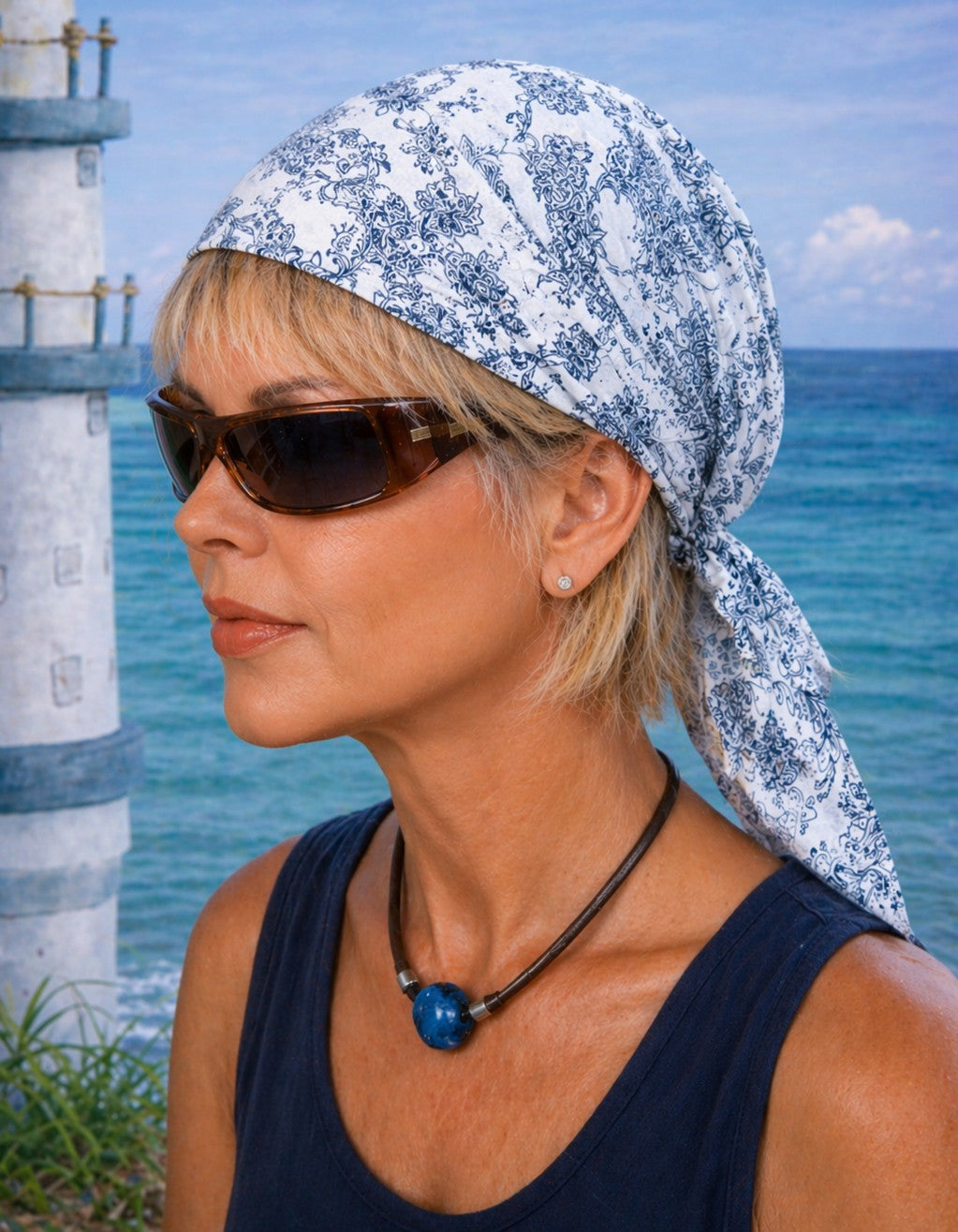 Reife Frau mit blondem Haar trägt ein fertig gebundenes Bandana-Tuch hinter dem Ohr und eine Sonnenbrille vor maritimem Hintergrund mit Meer und Leuchtturm