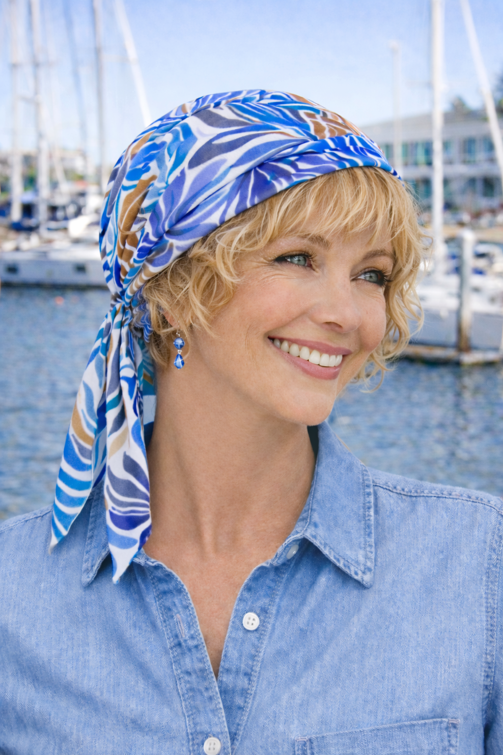 Frau mit blau gemustertem Chemotuch und blonden lockigen Haaren darunter lächelt am Yachthafen und trägt eine helle Jeansbluse