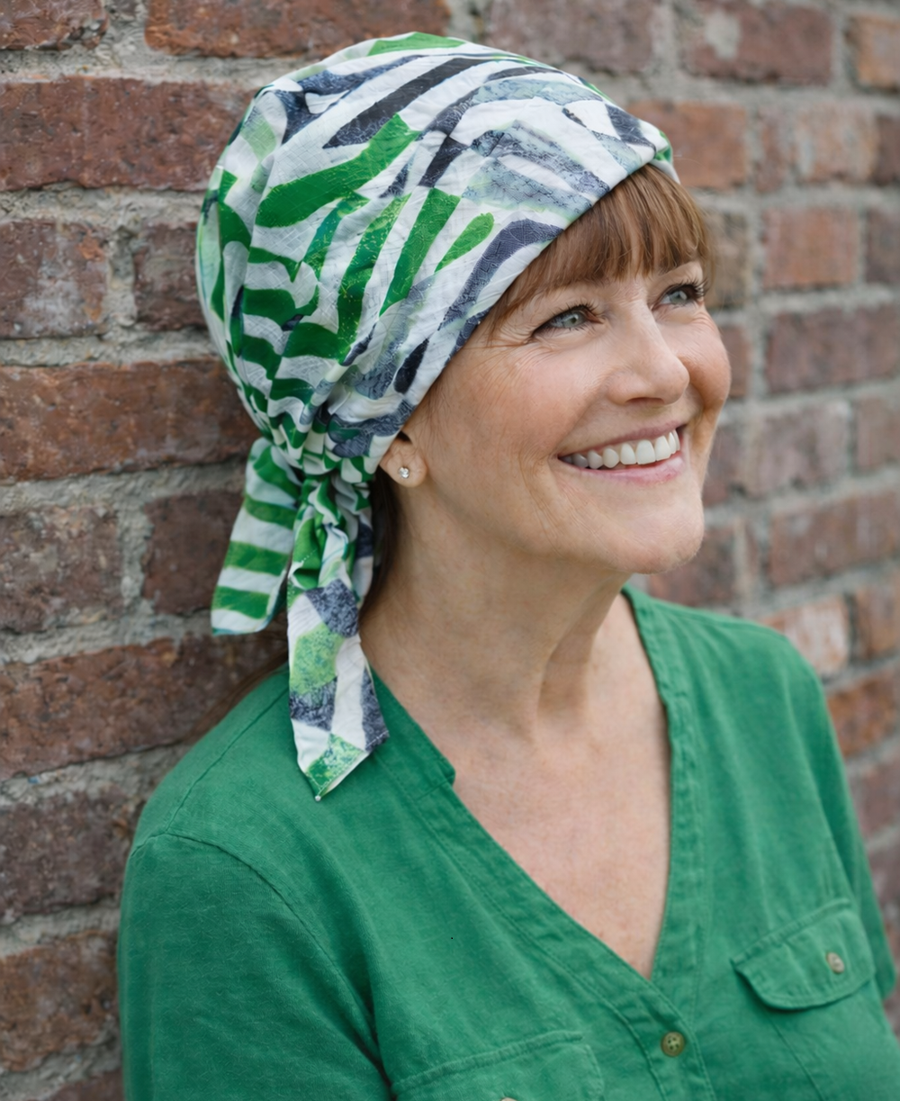 „Reife grauhaarige Frau trägt grün gemustertes Chemotuch aus leichter Baumwolle ohne Binden, sitzt entspannt im Strandkorb am Meer.“