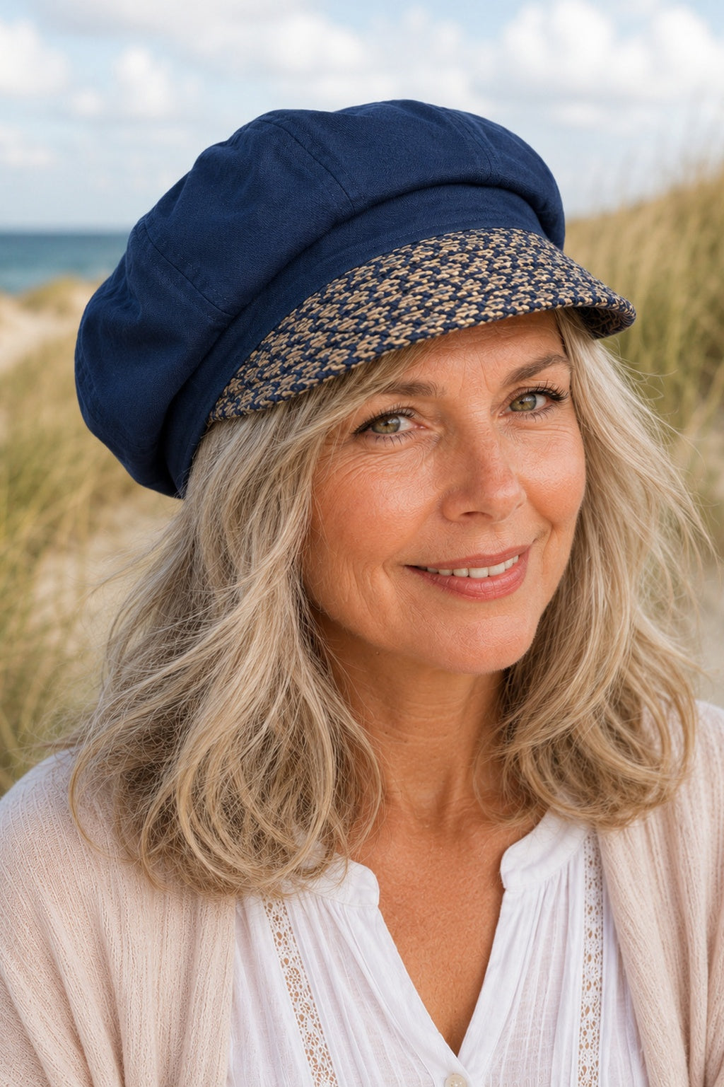 ältere Frau mit grauen Haaren trägt eine dunkelblaue Damen Schirmmütze aus Leinen und Baumwolle am Strand mit Dünen im Hintergrund.