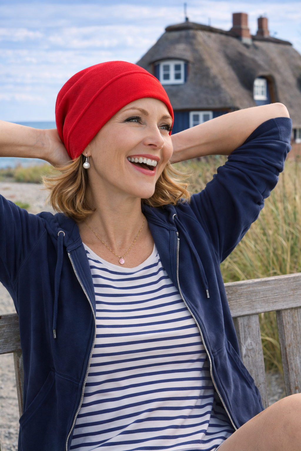 Lachende Frau mit mohnroter Sommermütze aus Baumwolle sitzt entspannt auf einer Bank am Meer, maritimer Hintergrund mit Reetdachhaus