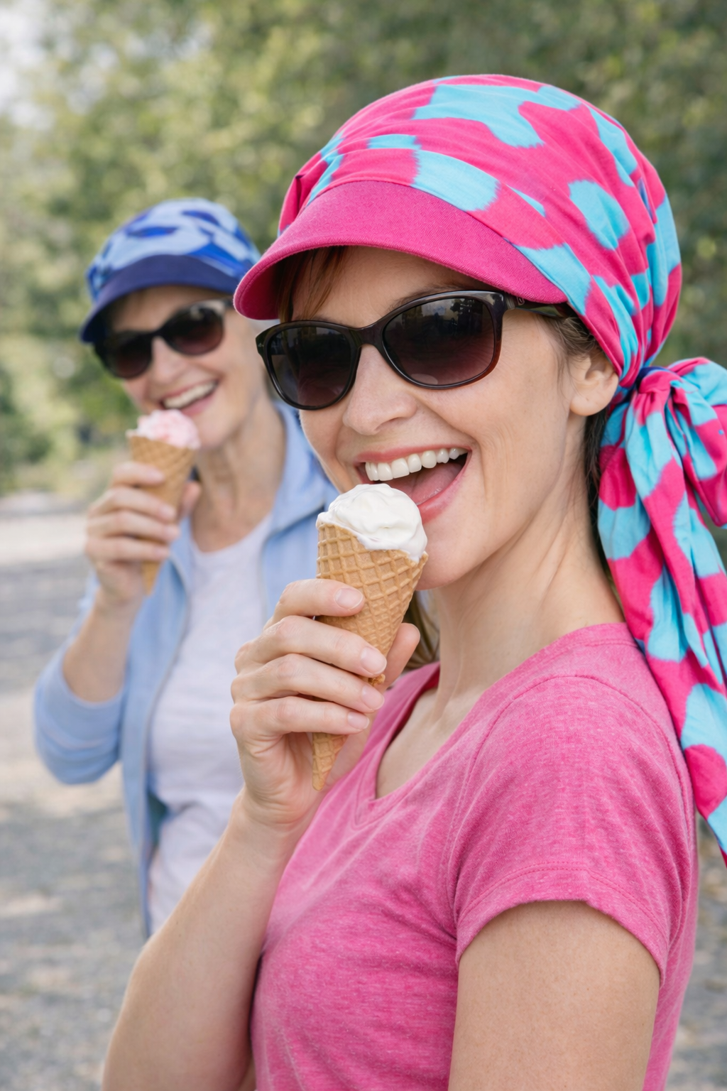 Zwei Frauen mit Sommer Kopftüchern mit Schirm und Sonnenbrillen essen Eis im Freien.